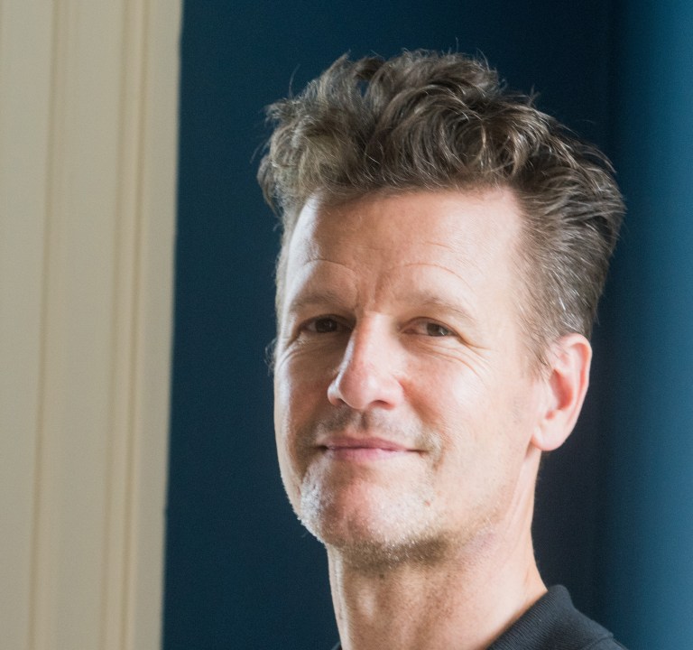 A closeup headshot of Geoff Nuttall over a blue and white background.