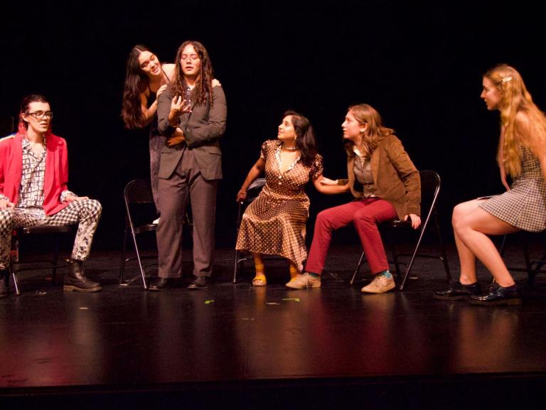 Six performers in varying outfits (a brown dress, a red jacket with checkerboard pants, etc.) sit and stand on stage. Illuminated by stage light, they all gaze engagedly at the two standing performers at the middle left.