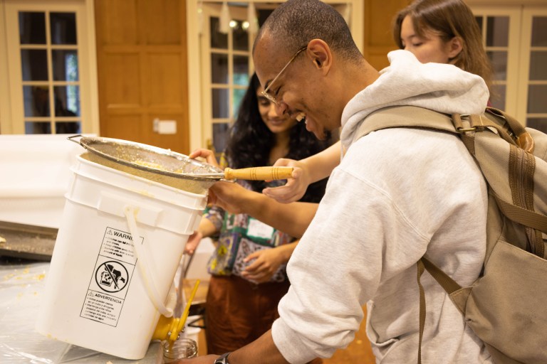 Jadon Geathers tilting a white bucket with a sieve on top, pouring honey into a glass jar through a spigot.