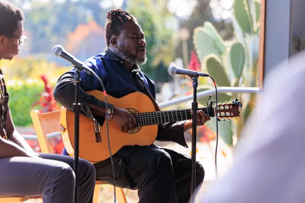 two Black individuals sit side-by-side in chairs in front of microphones. the person on the right, who is the main focus of the photograph, strums a guitar and sings with his eyes closed