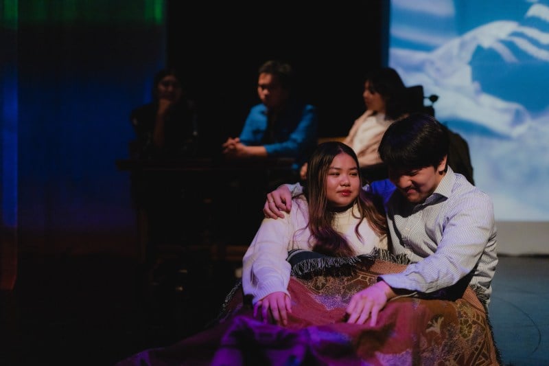 On a darkened stage, three figures sit at a table in the back while a young couple cuddle in the front.