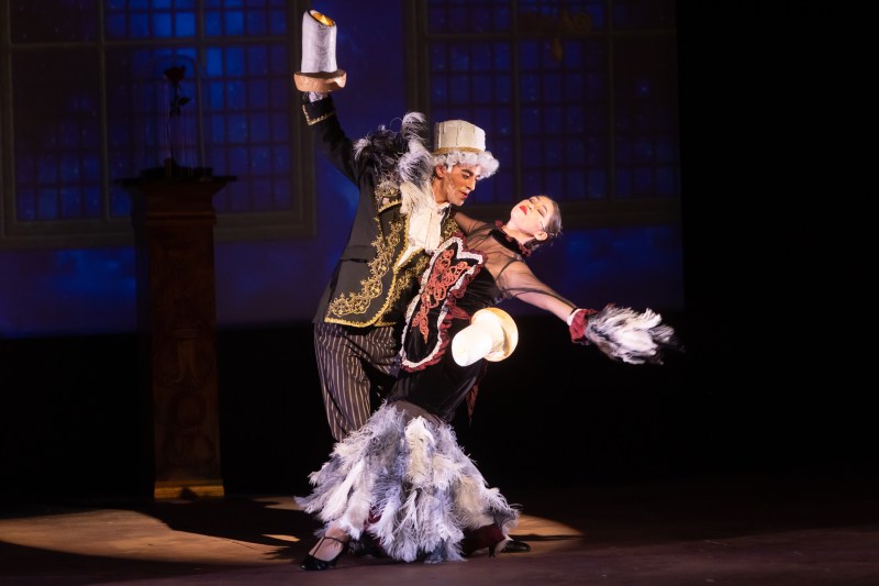 A man in traditional European costume and with candles extending from his arms tangoes with a lady dressed in feathers.