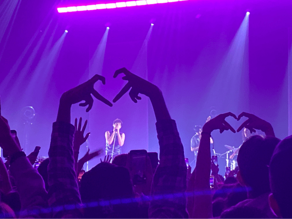 a man stands on a stage singing into a microphone; two members of the crowd make finger hearts toward him