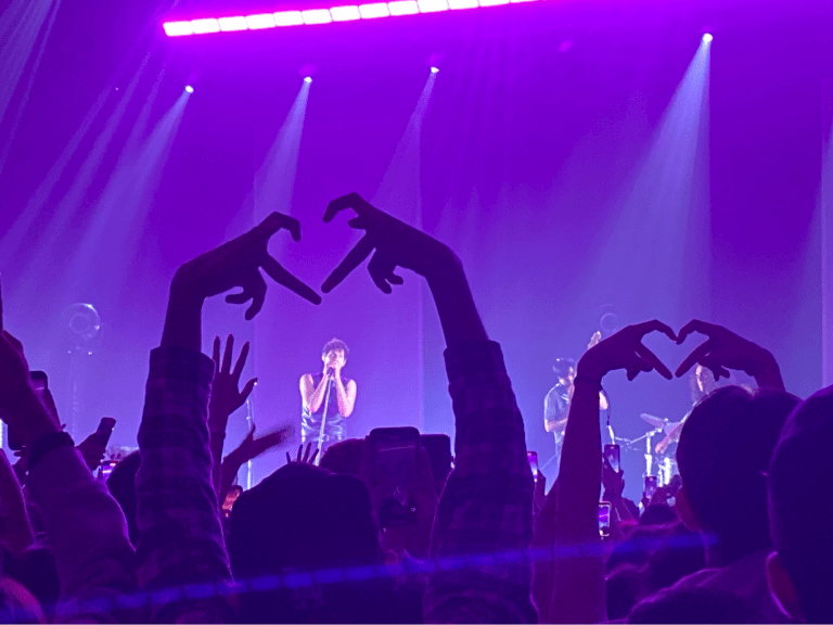 a man stands on a stage singing into a microphone; two members of the crowd make finger hearts toward him