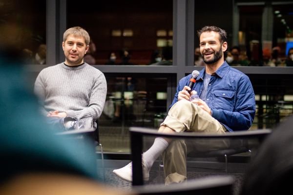 Person in gray sweater sits next to person in blue jacket holding a microphone.