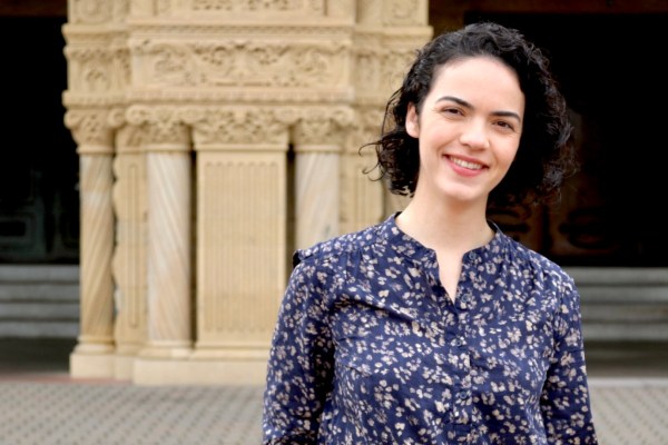 a person with light skin and dark brown hair smiles and stands in front of a classic stone pillar, wearing a dark blue blouse with white flower designs