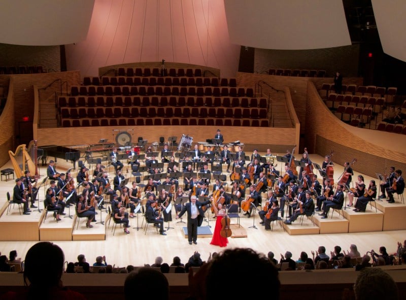 a group of musicians sits on a stage in four rows arranged around a conductor in a suit and a string soloist in a red dress