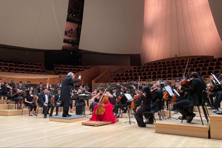 musicians in black suits sit in four rows with the conductor in the center and a cello soloist in a red dress next to him