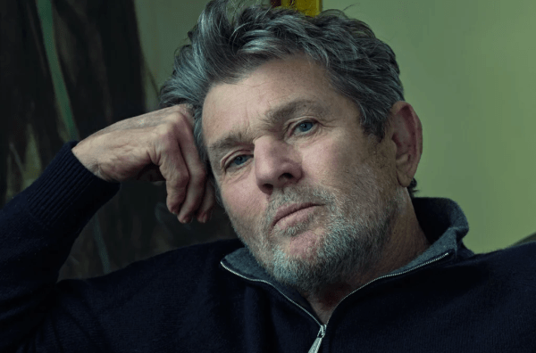 a photo of a white masculine person with gray hair and stubble, with their head leaned on their hand. the photo has dim lighting