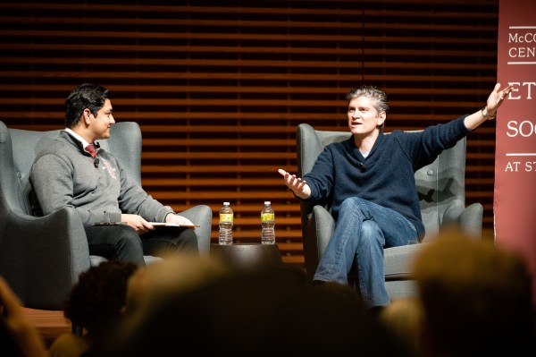 a photo of two people on seated a stage. the person on the left has tan skin and short black hair, and is wearing a gray sweater vest. the person on the right is pale with gray hair, is wearing a blue collared shirt and jeans, and is gesturing with an arm outstretched.