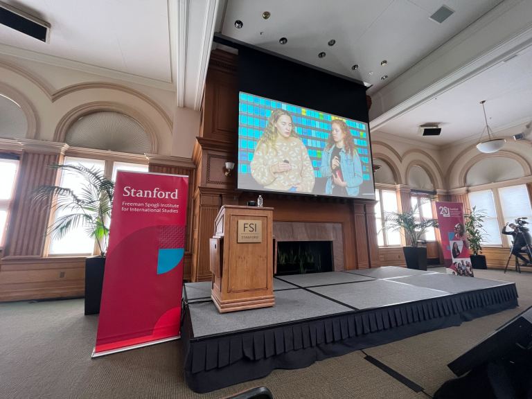 Two speakers on zoom are projected on a screen behind a podium at the Freeman Spogli event.