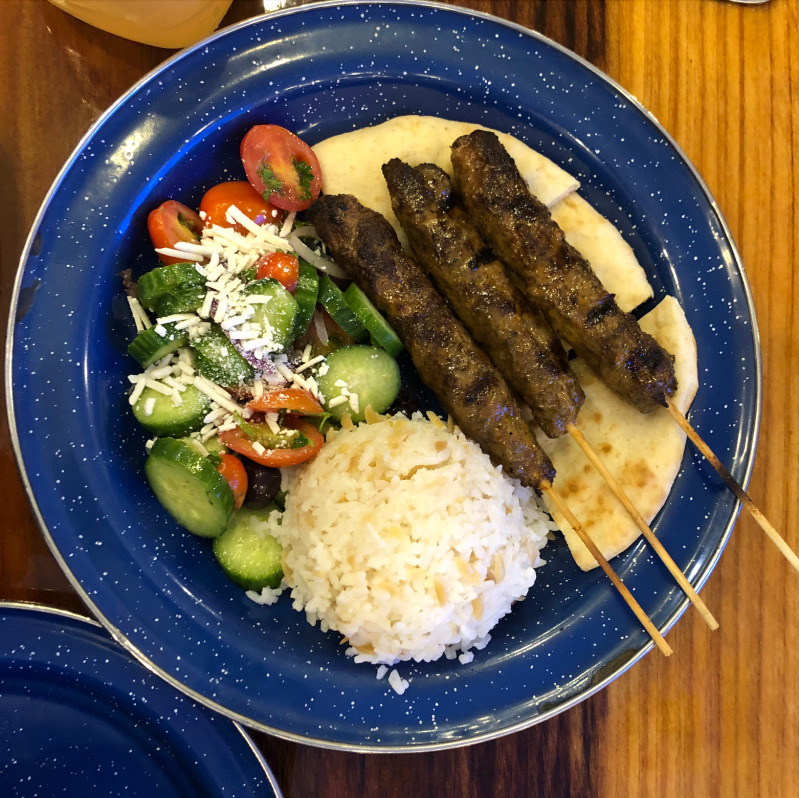 Cucumber salad, white orzo and 3 beef skewers sit on top of a bed of pita bread on a speckled blue plate.