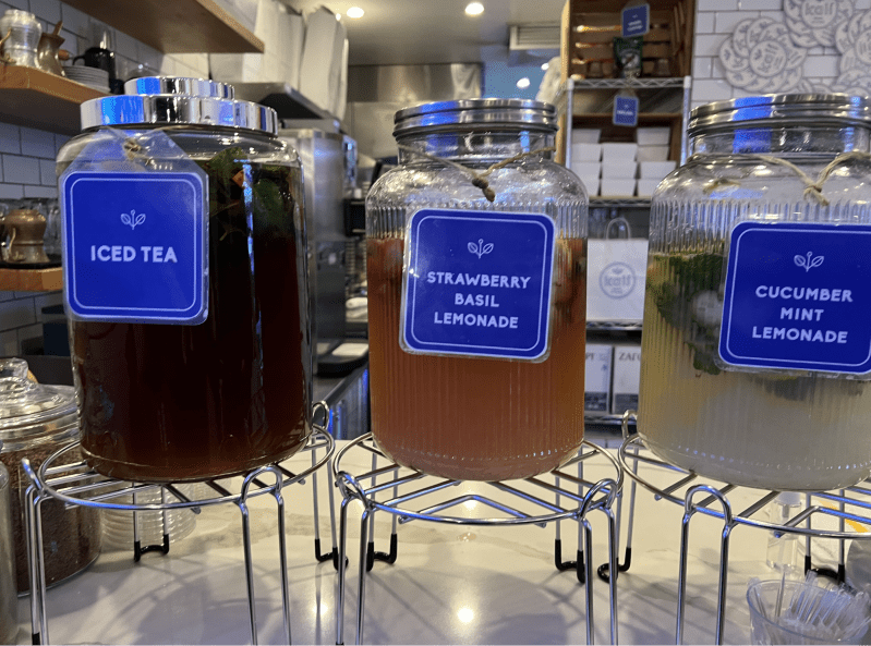 Three glass containers hold colorful drinks. The left drink is a dark brown iced tea. The center drink holds pink strawberry basil lemonade. The right drink holds light green cucumber mint lemonade.