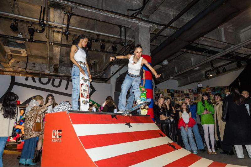 Models skateboard down red and white striped skate ramp wearing white tank tops and jeans. 