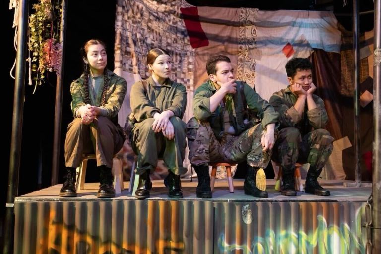 Four actors in green costumes sit on stage looking at something beyond the camera.