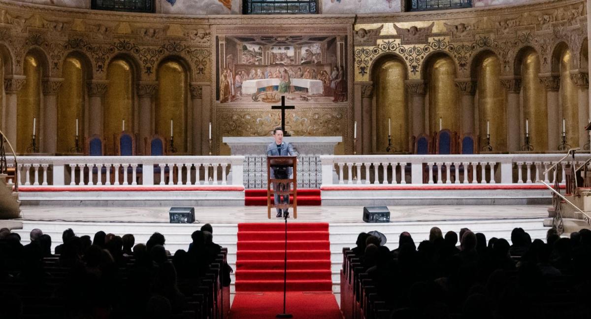 A writer standing in Memorial Church, with rows of audience sitting in front of him