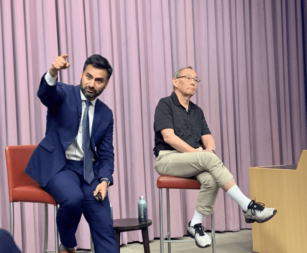 Two men sit on chairs with one man in a suit and the other in a tshirt and trousers.