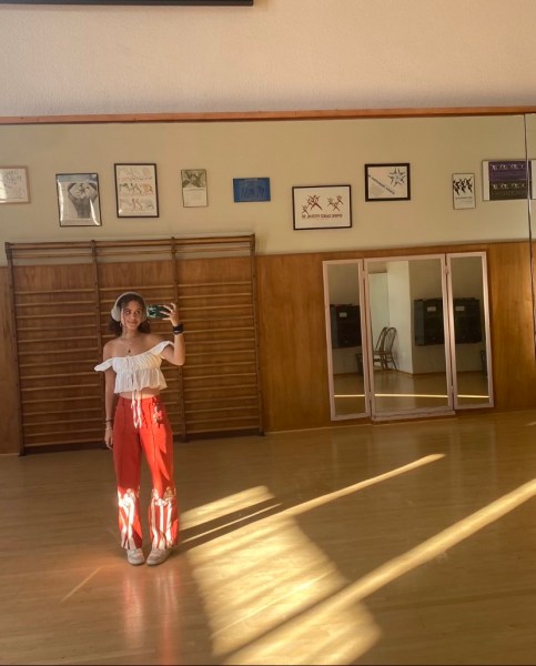 Olivia Jessner stands in a light wood room wearing a ruffled, off-the-shoulder white top and orange pants with white mushroom growing from the bottom.