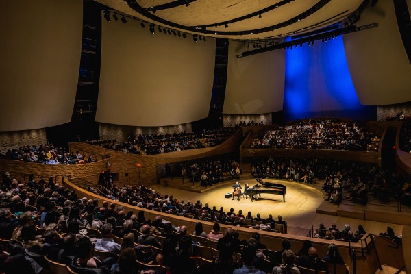 a photo of a large crowd sitting in a concert hall around a piano player and cellist, whom are spotlighted on stage