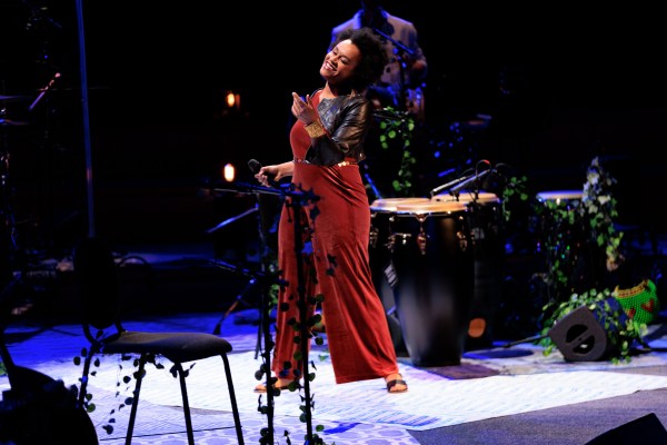 a person with brown skin and short kinky black hair stands smiling on a stage, which is decorated with green plastic vines