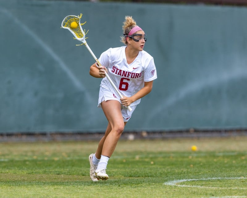 A womans lacrosse player runs along the field with possession of the ball