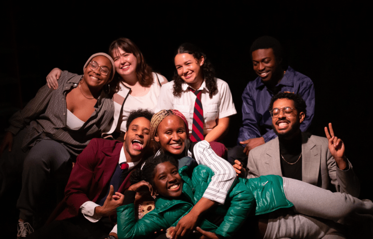 Against a dark backdrop, eight actors and directors smile at the camera and hug each other.