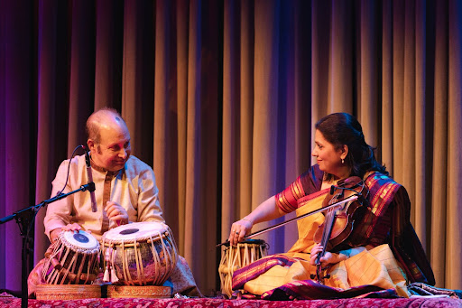 Kala Ramnath is seen playing violin. Abhijit Banerjee sits to her right on a drum.