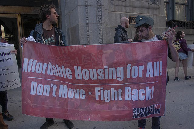 two men holding a sign that says, "Affordable Housing for All. Don't Move -- Fight Back"