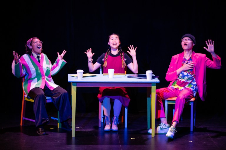 Three actors sit around a table with their arms up, facing the audience of TAPS show, "CS106A."