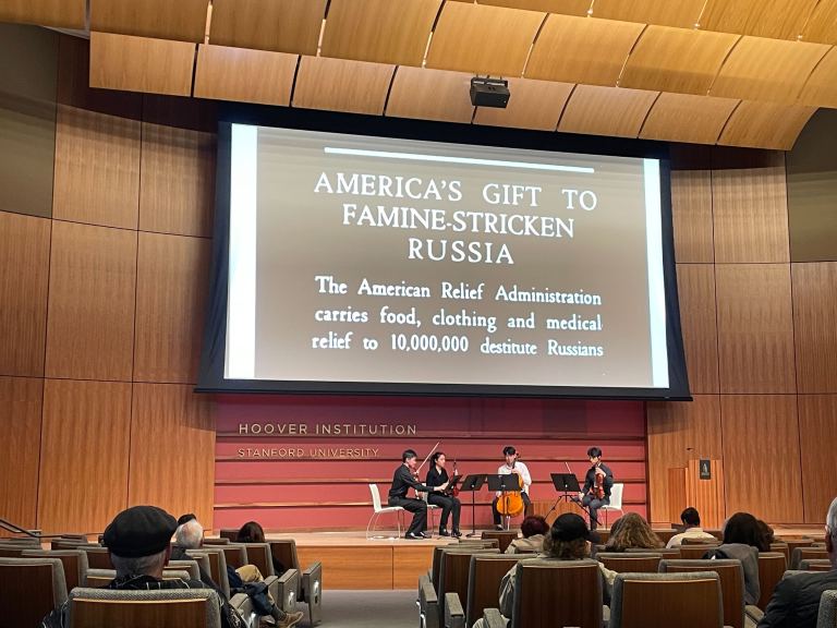 A photo of the Hauck Auditorium of Traitel building, with a documentary projected. Four musicians are playing on stage.