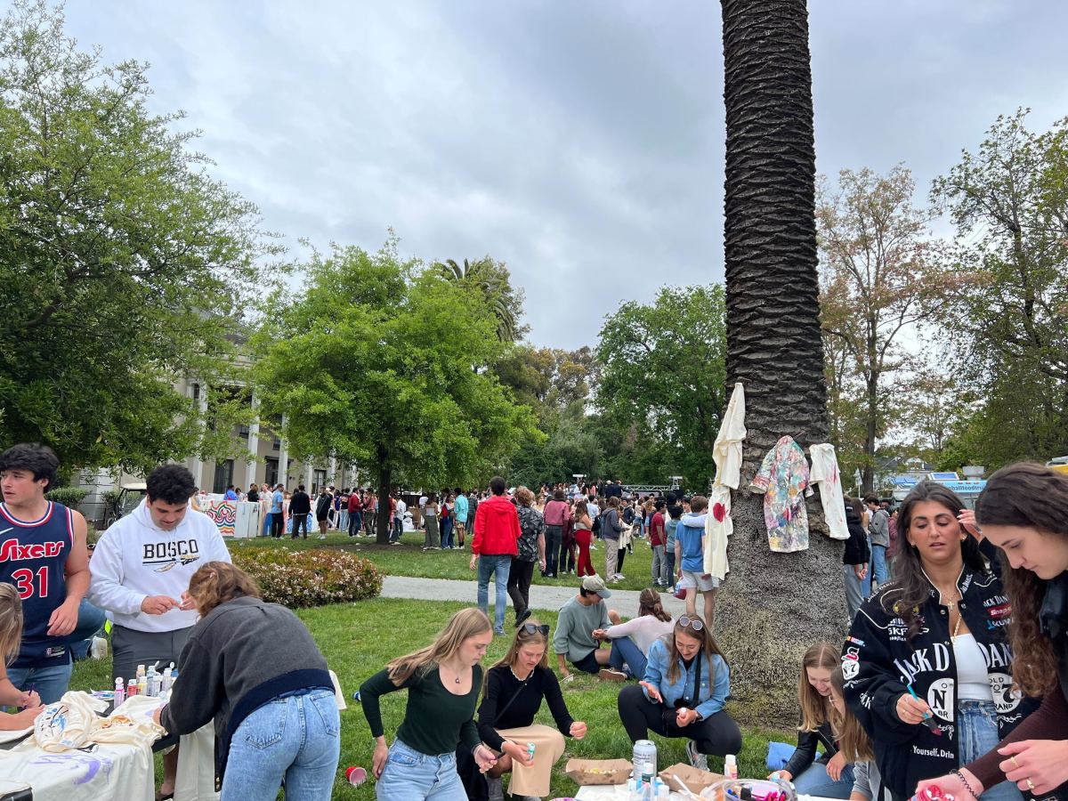 Students decorate shirts at the base of a tree while others gather in front of the Sigma Nu house behind the tree.