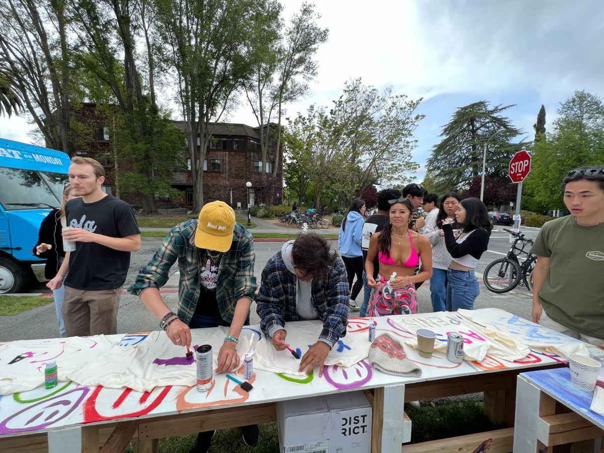 Three students paint on shirts on a shared long table.