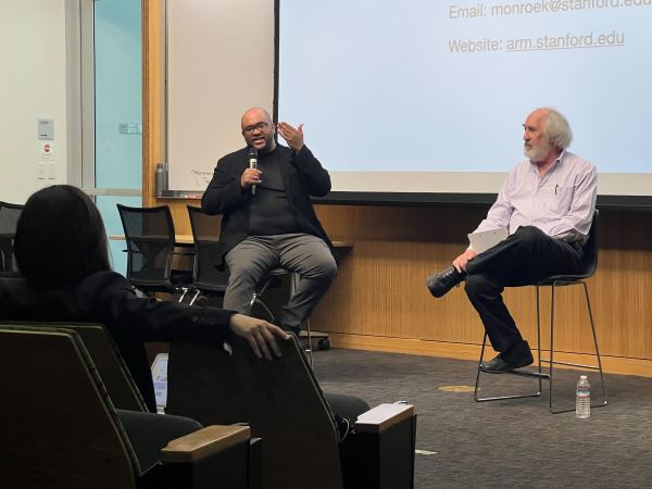 Two male speakers sit in front of a lecture hall, with a projected white slide behind them containing a link to a website.