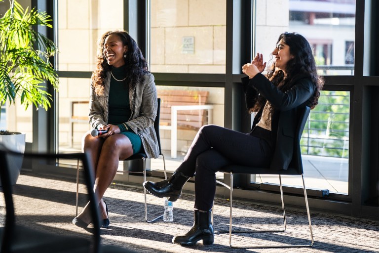 Stegner Fellows Christell Victoria Roach (above, left) and Rabia Saeed (right) sit next to each others in chairs. they are in front of the large windows of the Mackenzie room, lit by the natural light of the evening.