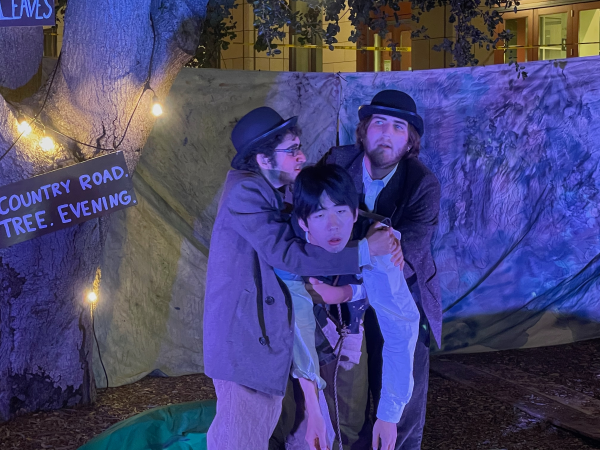 Three actors huddled around the middle of a set. The actor in the middle is hunched over, and held up on both sides by his fellow actors.