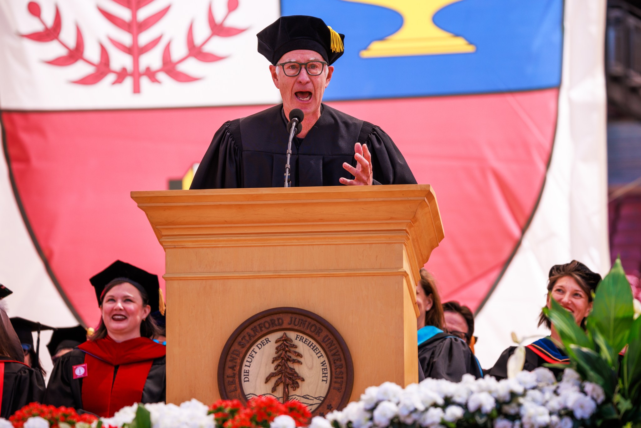 John McEnore wears a black cap and gown. He is standing at a podium as he speaks to a crowd of graduating students.