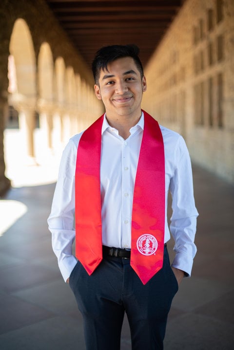 Richard stands between the arcades at Main Quad. He is wearing a white dress shirt and dark pants. He has a red sash around his neck.