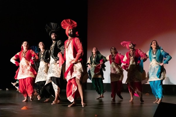 a photograph of eight people dressed in ornate uniforms colored red, black or blue; they pose on a stage with their hands on their hips and grinning facial expressions, looking to the upper right corner of the room behind the camera