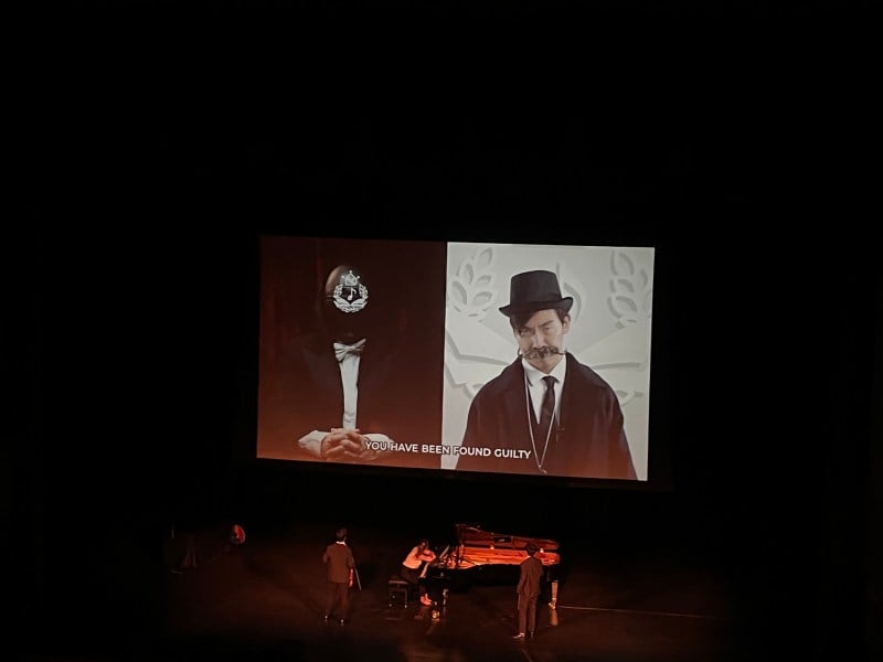 The left side of a large screen depicts adjudicator of TwoSet Violin-imagined "Classical Music Academy" accusing TwoSet of infringing upon copyright, while the right side shows a defeated-looking Eddy Chen. Chen, TwoSet duo partner Brett Yang and Sophie Druml watch the screen from below.