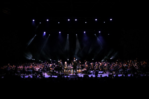 A five-person rock band is center stage, surrounded by a 115-person orchestra dressed in black. The scene is dark and dramatic.