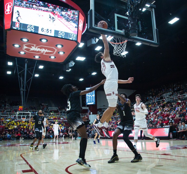 A basketball player mid air about to dunk a ball into the net. A few players gather around him.