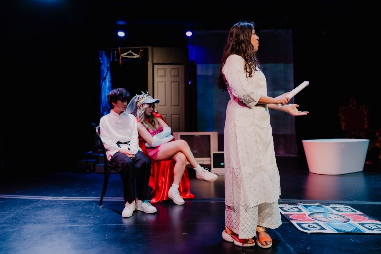 a photo of a stage, with two people sitting on chairs in the left-side background; the one on the left is wearing a white button-up shirt and black pants, and is sitting ridigly, while the person on the right is leaning casually and wearing a bright red cape. in the foreground to their right, a person with long dark hair and a long white kurti-like tunic is in the middle of speaking.