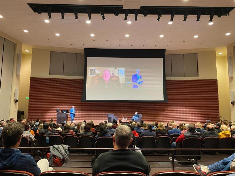 Packed auditorium with speakers on stage and Zoom participants on a projector