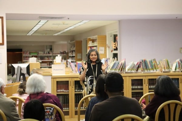 Storyteller Roopa Mohan talks to an audience in a library with shelves stocked full of books behind her.