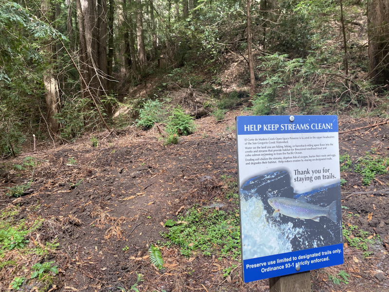 A sign that reads "help keep streams clean" and "thank you for staying on trials" with trees and bushes in the background.