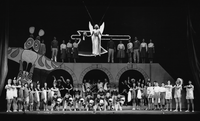 Dozens of Gaieties performers onstage, dressed as football players, Native Americans, and cheerleaders. A woman in an angel costume is suspended from the ceiling.