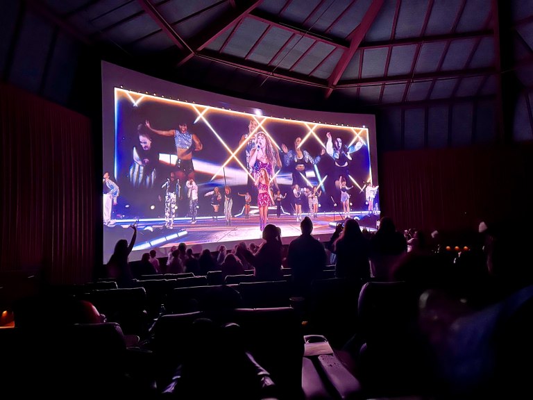 Silhouettes of audience members dancing before a screen playing "The Eras Tour Concert Film" inside a dark theater.