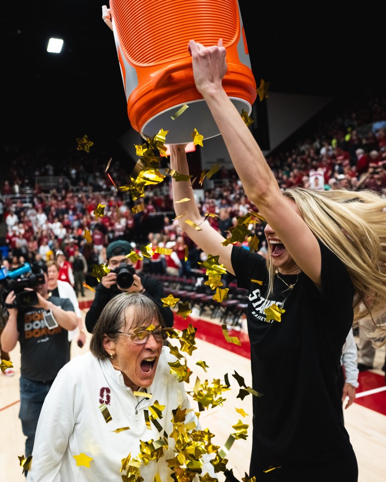 A woman dumps an orange bucket of gold confetti onto a shorter woman, who reacts in shock