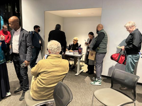 People standing around Clarence B. Jones sitting at a desk, waiting for him to sign copies of his book.