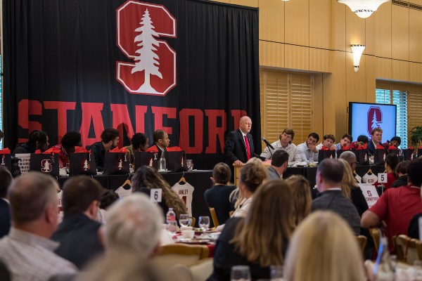 A man stands at a podium next to a row of sitting Stanford football players, in front of a crowd of listeners